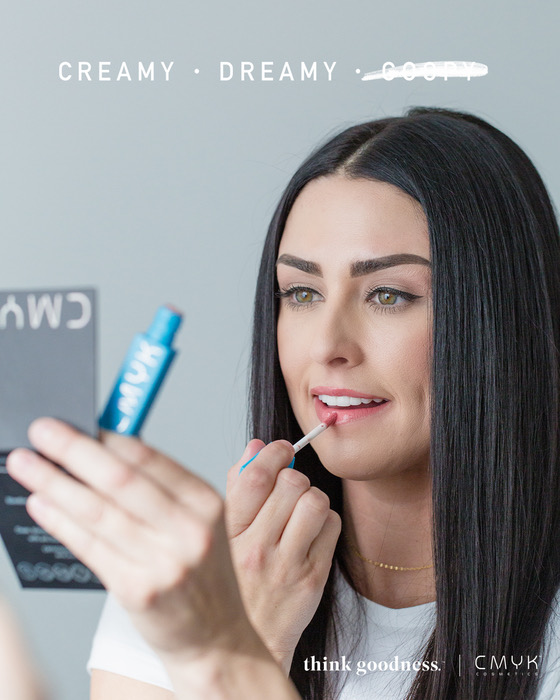 Woman holding compact mirror putting on CMYK Cosmetics lip gloss