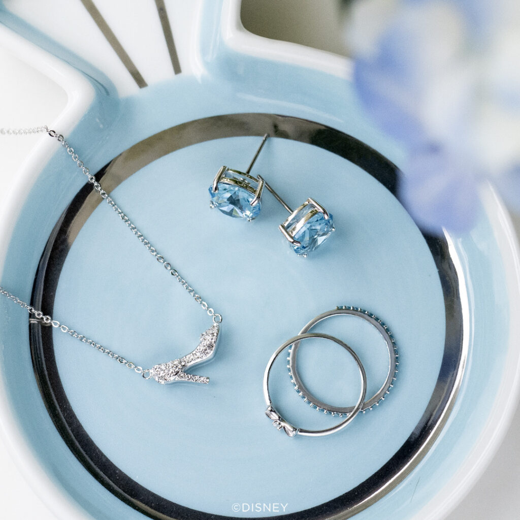 A picture with a blue background with Disney Cinderella jewelry blue Clara stud earrings, pave slipper necklace and stackable rings that has kind wrote on one ring 