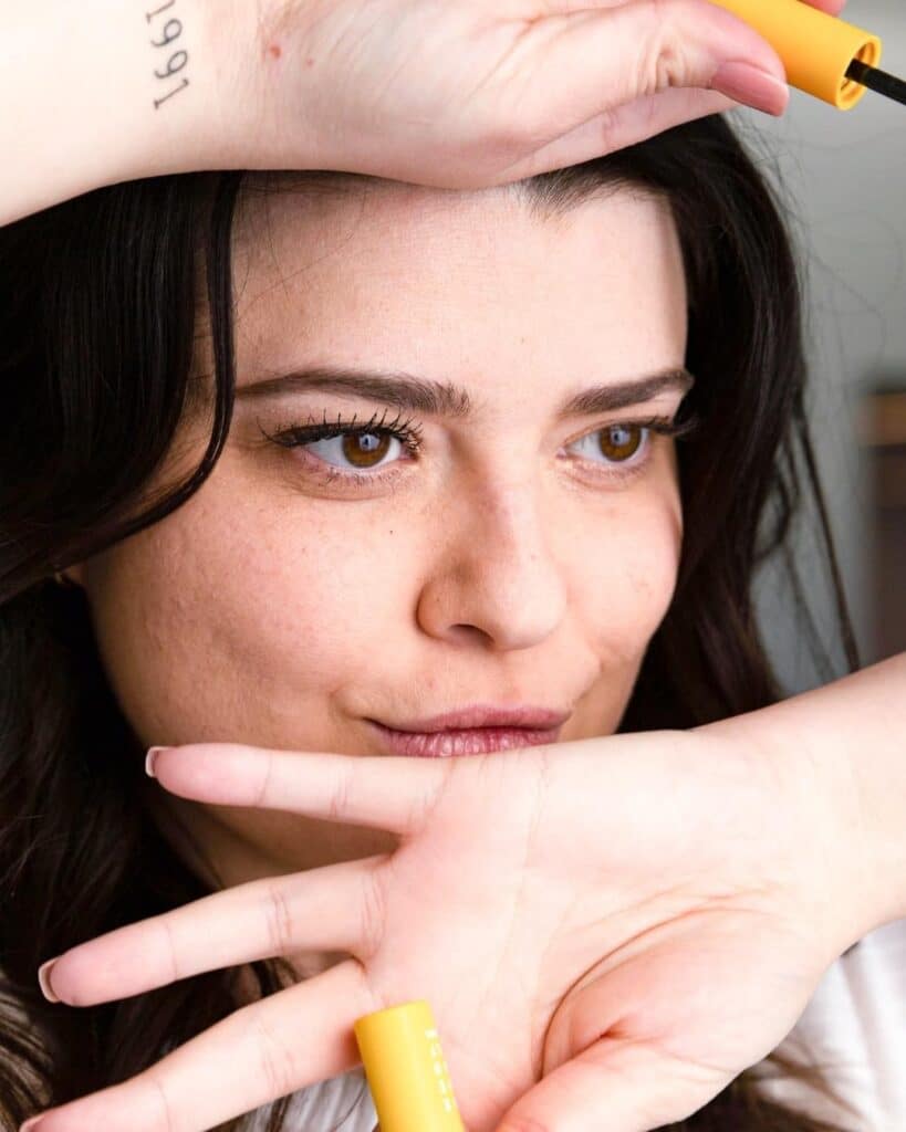 An image of a model with dark hair wearing the CMYK mascara and holding the mascara 