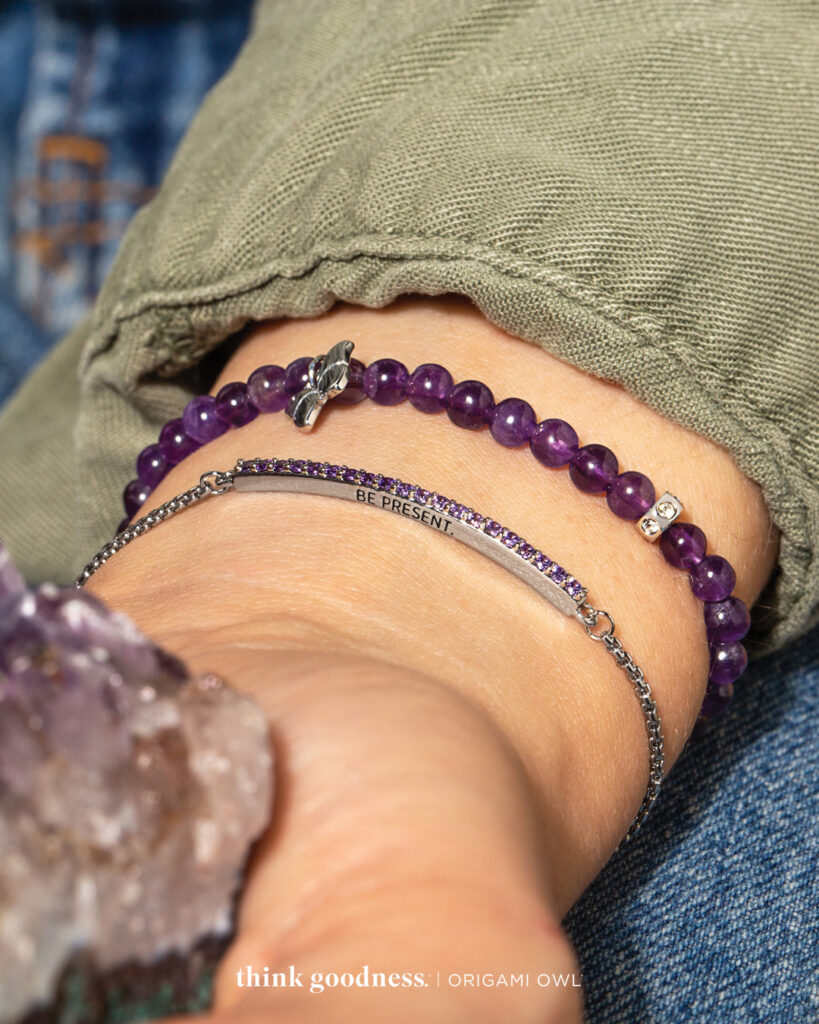 a womans wrist showing a purple beaded stretch bracelet and a silver bolo bracelet with purple crystals that says be present