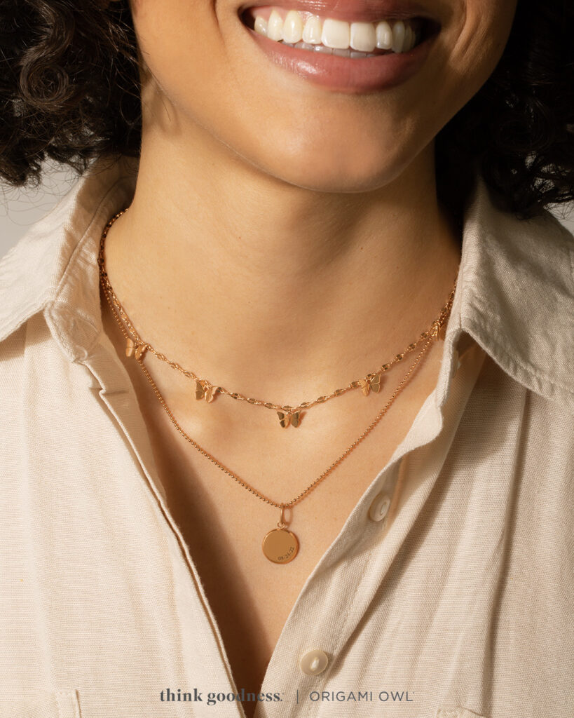 A woman’s neck in a beige blous wearing a rose gold butterfly necklace and a rose gold round inscriptions necklace