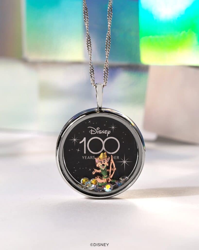 A holographic background with a silver locket and chain. In the origami owl locket are the Disney 100 plate and Disney 100 Robin Hood charm