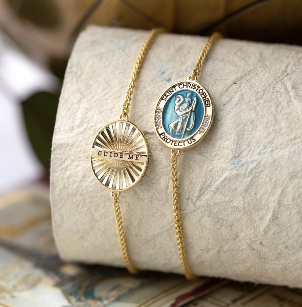 a beige round jewelry display showing one side of the saint christopher medal with image and protect us and the other side that says guide me in gold 