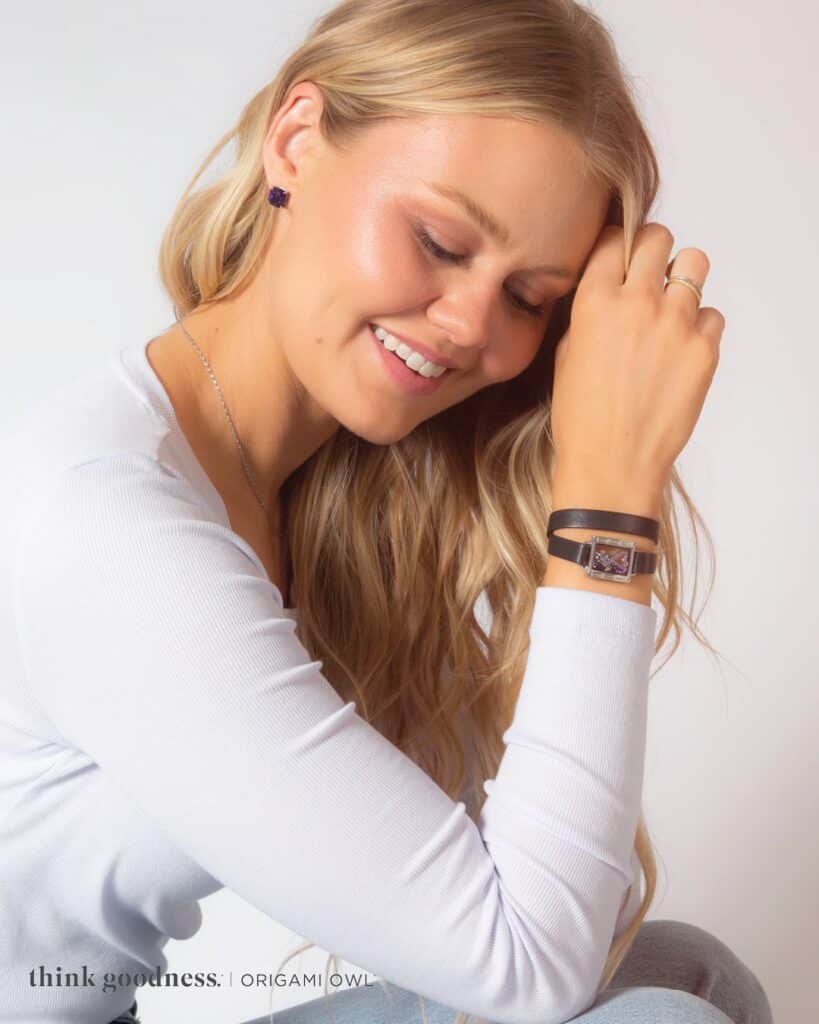 Bella is sitting sideways with her elbows on knees wearing a white long sleeve shirt wearing the origami Owl locket wrap bracelet, earrings and necklace