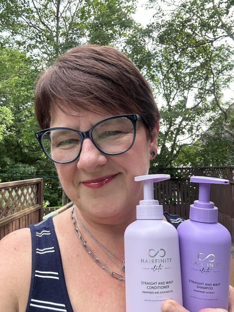 A picture of Colleen Evans holding Hairfinity Elite Shampoo and Conditioner with trees in the background