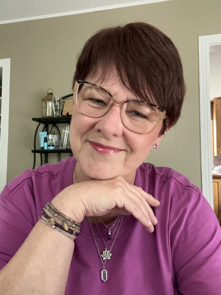 an image of Colleen Evans in a purple t-shirt wearing layers of bracelets, necklaces and earrings. My arm is under my chin showing my confidence and empowerment. I am wearing Willing Beauty Skincare, CMYK Cosmetics which are all clean products