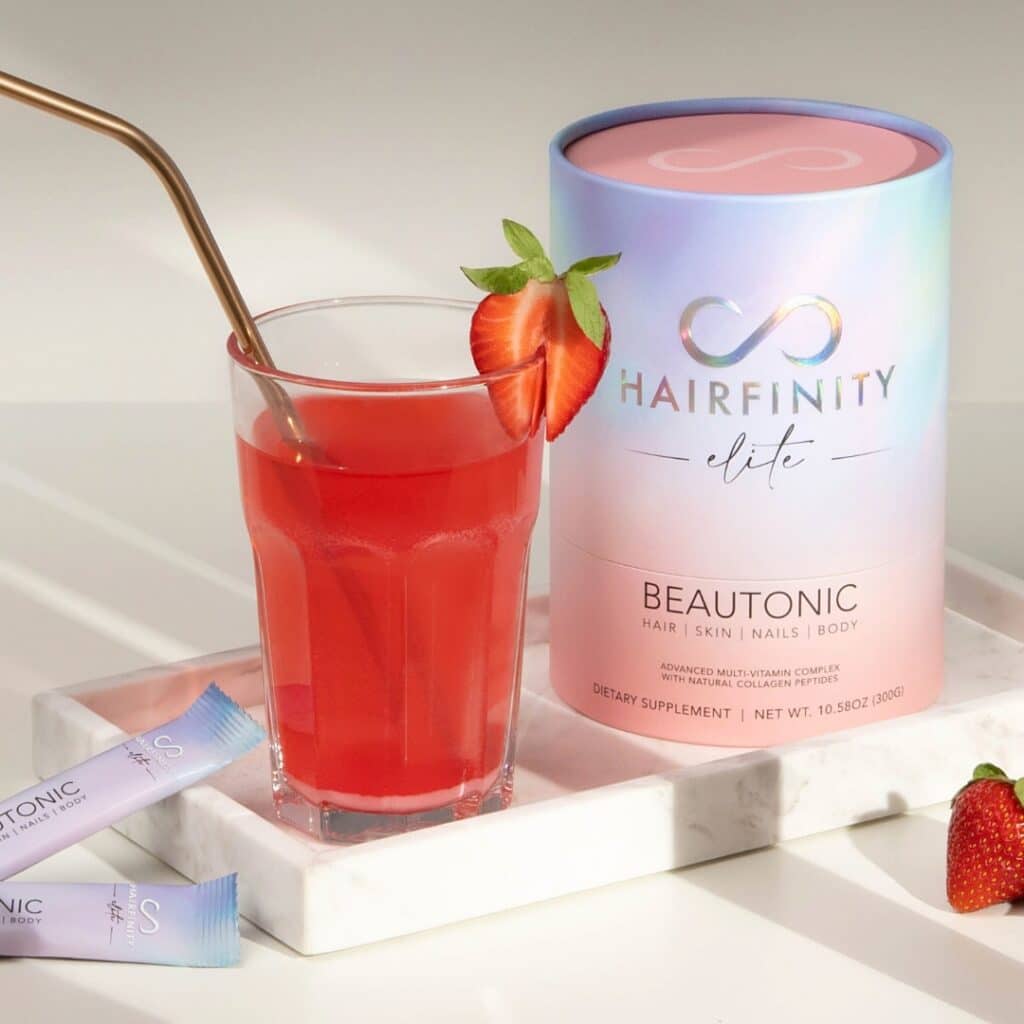 a light image with white dishes showing a container of hairfinity elite beautonic and a glass of beatonic. It's a red liquid with a straw in the glass and half a strawberry on the rim