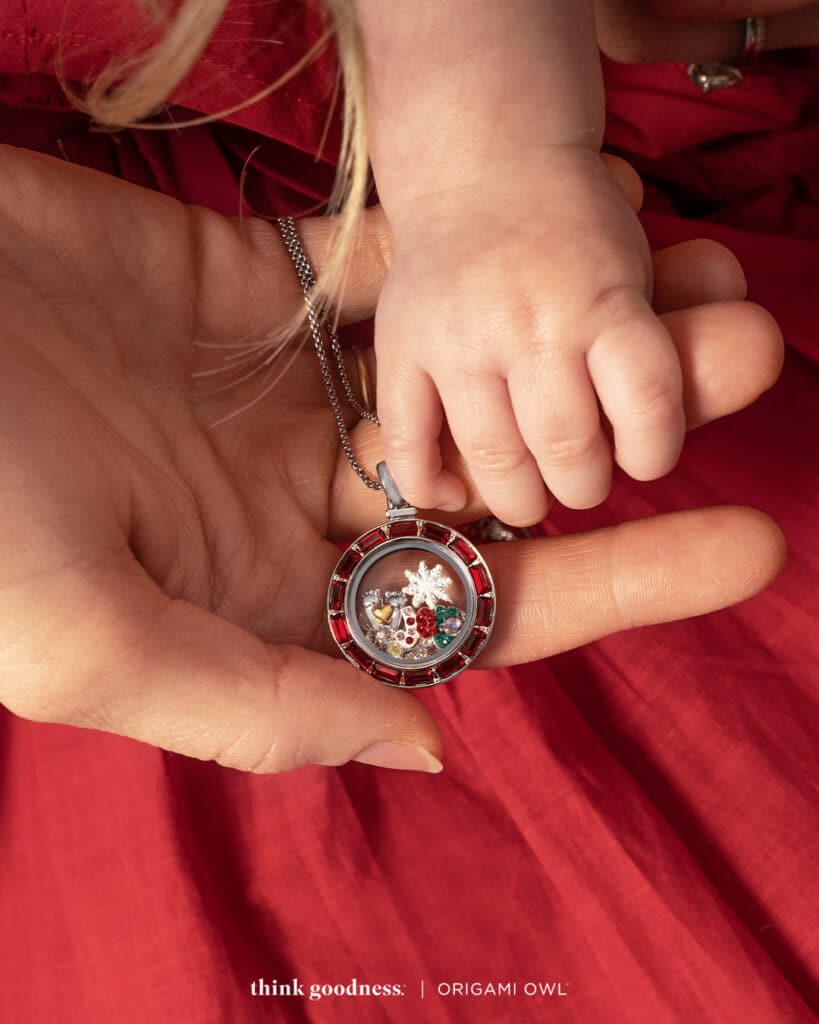 a red background with a persons hand and a babys hand holding a siam crystal baguette locket with holiday charms
