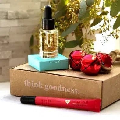 a think Goodness corrugated box with a willing beauty peppermint lip oil laying in front, a bottle of willing beauty born to glow skin elixir on top of the box next to 3 christmas bells with greenery in the background